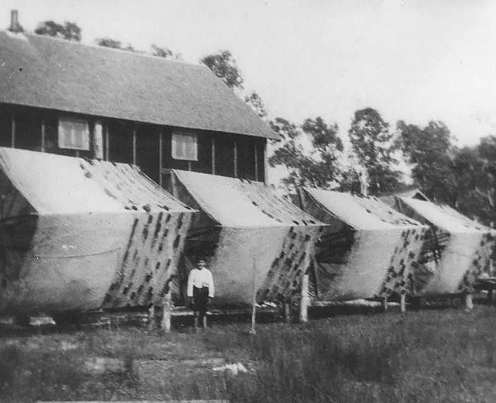 A row of canvas tents stands in front of a large wooden building, with one person in the foreground.