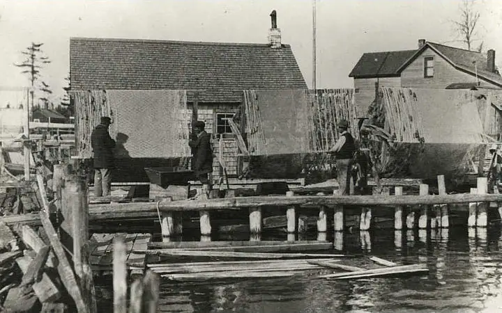 Black-and-white waterfront photograph showing people working on a wooden pier beside waterfront buildings.