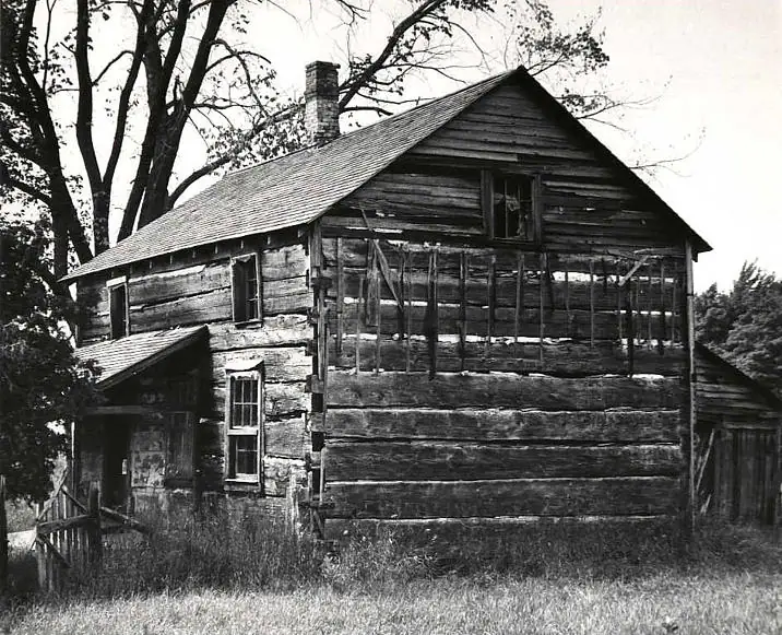 Weathered two-story log house with a gabled roof, chimney, porch, and tall grass around it.