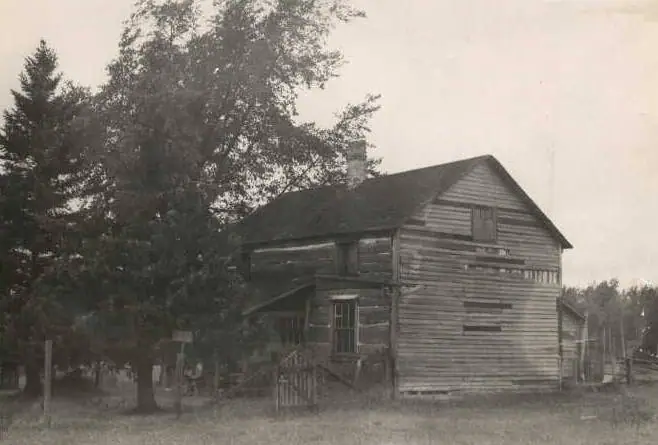 A weathered two-story wooden house stands among trees beside a fence and grassy yard.