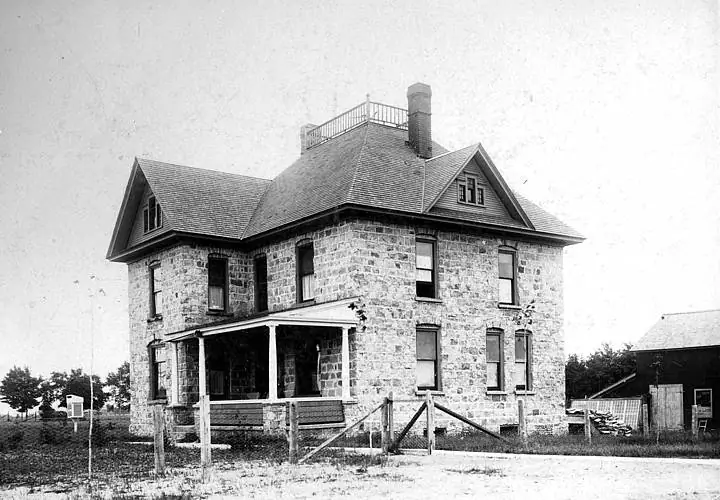 Black-and-white photograph of a large two-story stone house with a porch, fenced yard, and adjacent outbuilding.