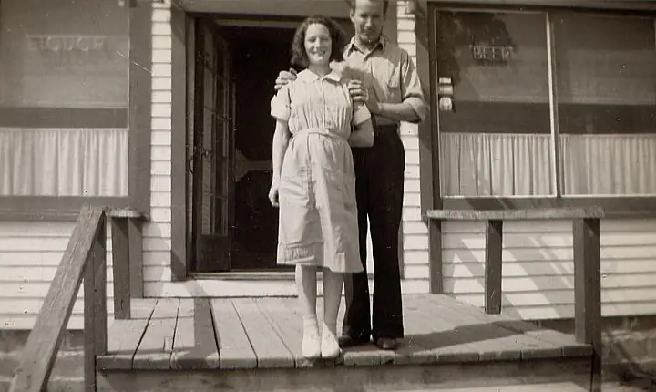 Two people stand together on a wooden porch outside a building with lettering on its windows.