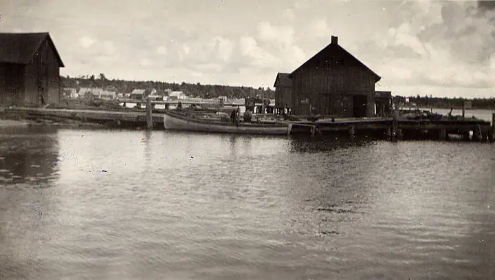 Black-and-white waterfront photograph showing wooden buildings, docks, a small boat, and dark clouds.