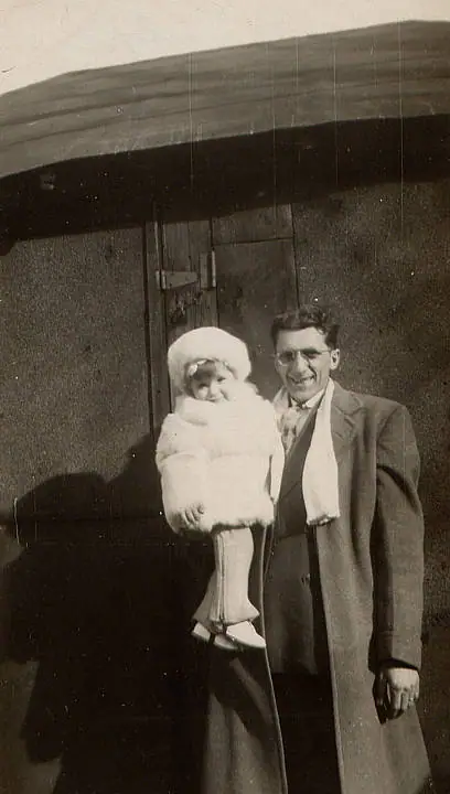 A man in a long coat holds a bundled child in front of a wooden doorway.