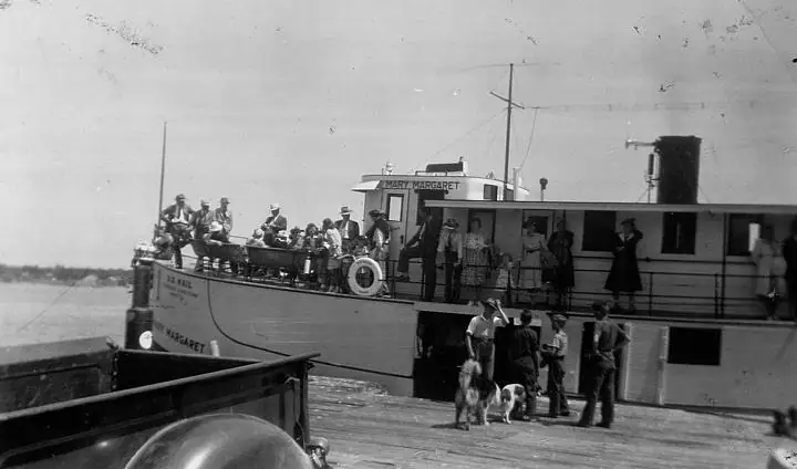 A ferry crowded with people is alongside a wooden pier, with a dog among people on the dock.