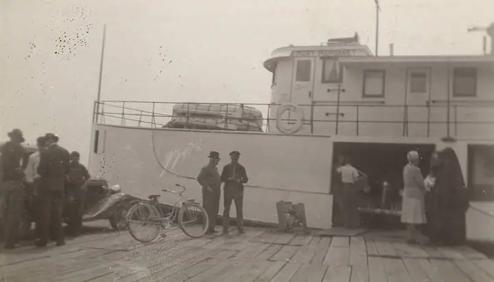 People gather beside a bicycle and car on the wooden deck of a ferry.