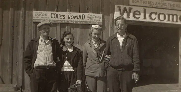 Four adults pose outside a wooden lodge beneath signs advertising lodging, fishing, and boat rides.
