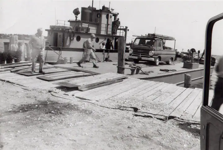 A workboat is moored beside a wooden dock while people and a van occupy the loading deck.