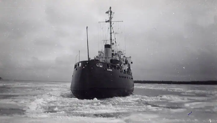 A working vessel pushes through broken ice under a cloudy sky.