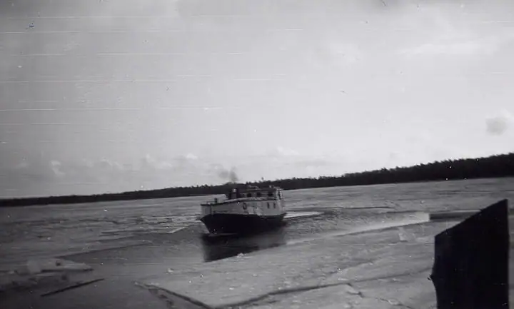 A small workboat moves through broken ice, with a wooded shoreline behind it.