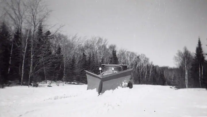 A snowplow truck pushes through deep snow at the edge of a wooded clearing.