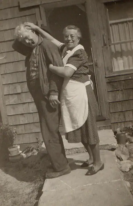 Two adults pose playfully outside a shingled house, with one holding the other's head.