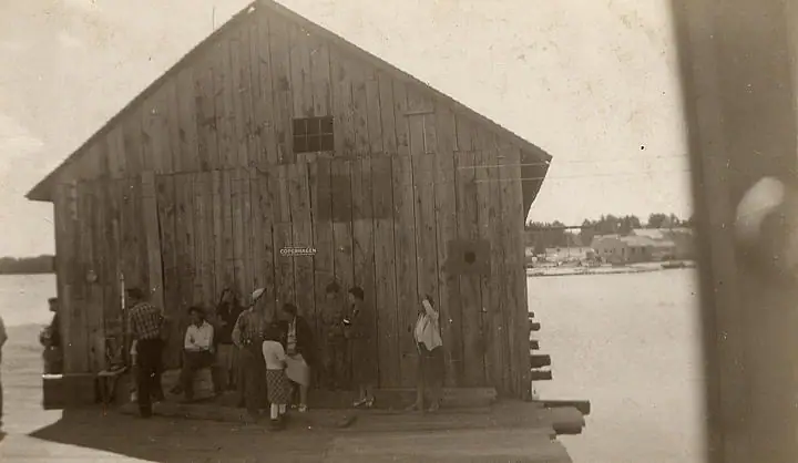 A group stands beside a large wooden building on a dock, marked with a small COPENHAGEN sign.