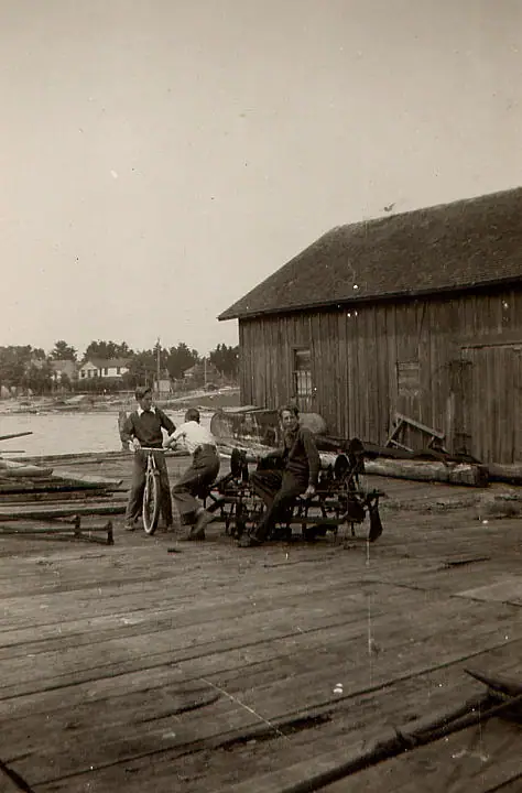 Three people gather beside a bicycle and wheeled cart on a wooden pier beside a large timber building.