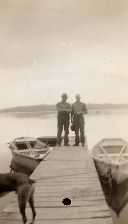 Two men stand on a wooden dock between two small boats, with a dog partly visible in the foreground.
