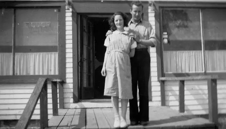 A woman and man stand together on a wooden porch outside a storefront with large windows.