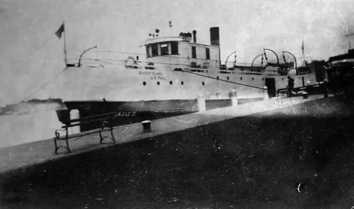 A large mail vessel is moored beside a dock, with lettering on its hull and a bench in the foreground.