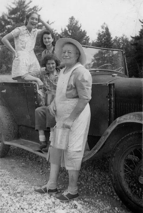 Four women pose beside a vintage open automobile, with one holding a metal pail.