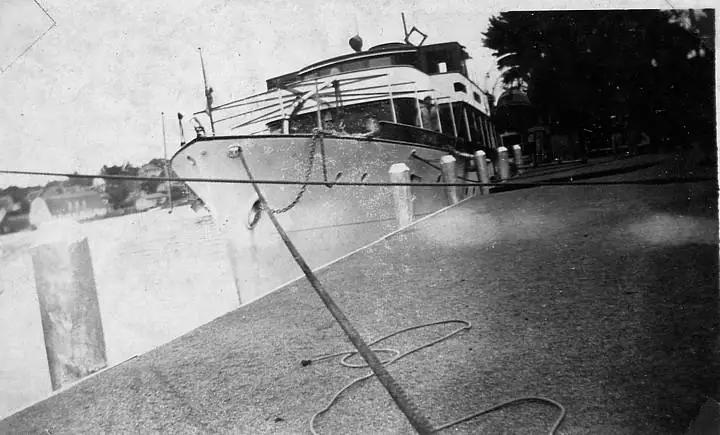 Black-and-white photograph of a passenger vessel moored beside a concrete pier, with a rope across the foreground.