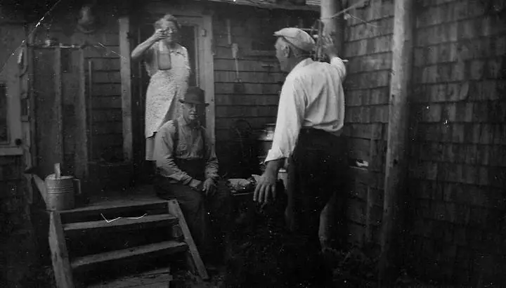 Three people gather beside a shingled building, with one woman holding a bottle near the doorway.