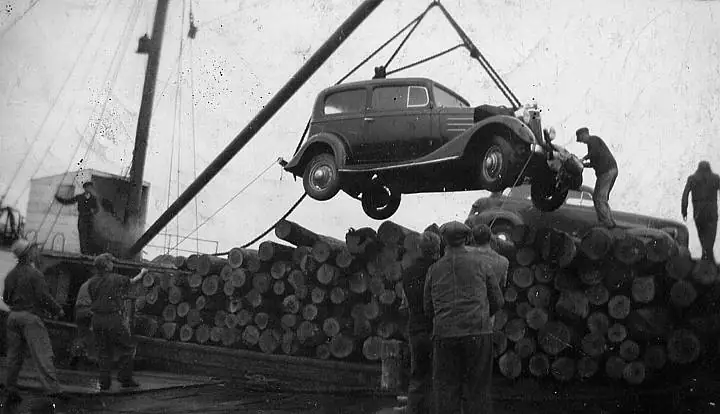 A car hangs from a crane above a deck loaded with logs while several men watch.