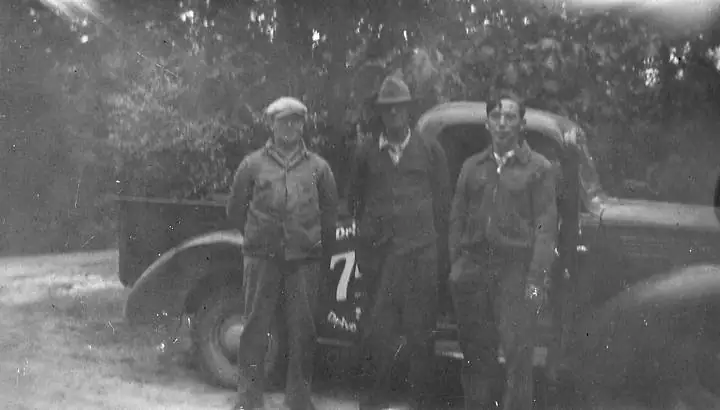 Three men pose beside a vintage truck marked with a large 7.