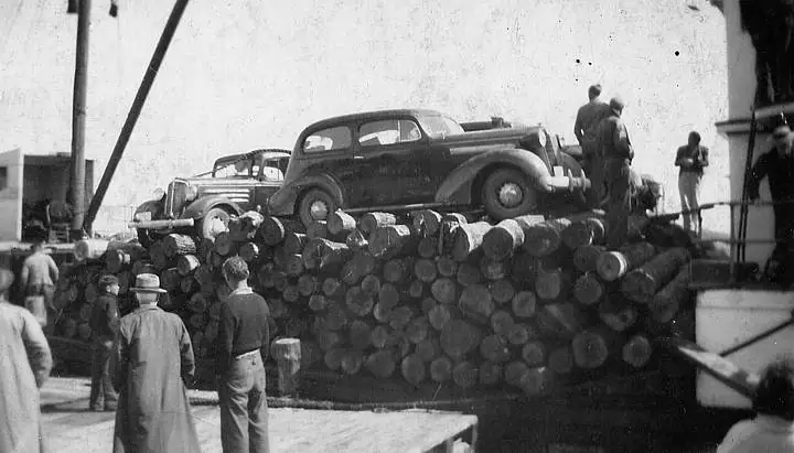 Two vintage automobiles sit atop a large stack of logs while people gather around it.