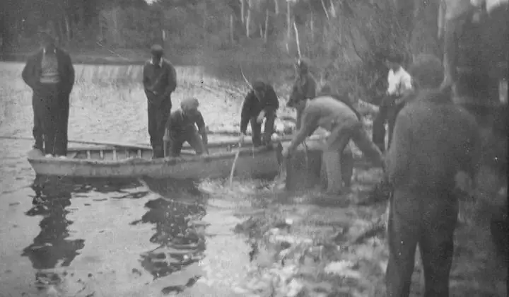 Men work beside a small boat at a wooded shoreline, with several people gathered in shallow water.
