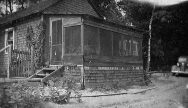 Black-and-white photograph of a small shingled house with a screened porch and a partially visible automobile.