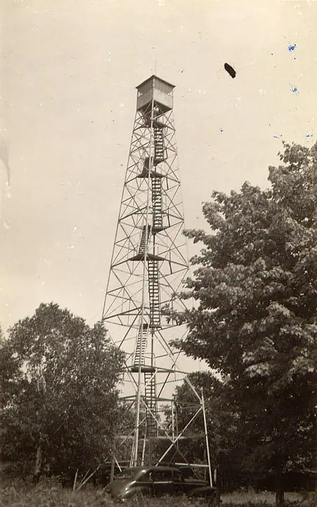 Lattice fire tower with multiple stair flights rises above trees; part of a car is visible at its base.