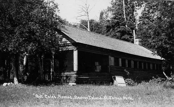 Black-and-white photograph of a long log cabin with a porch, surrounded by trees and tall grass.