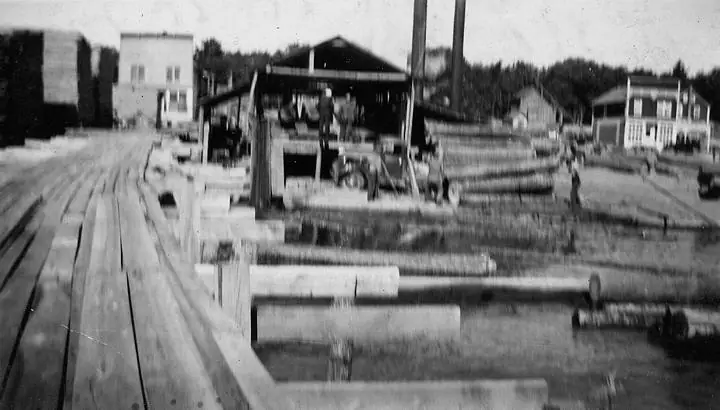 Black-and-white view of a sawmill yard with logs, machinery, buildings, and a plank walkway.