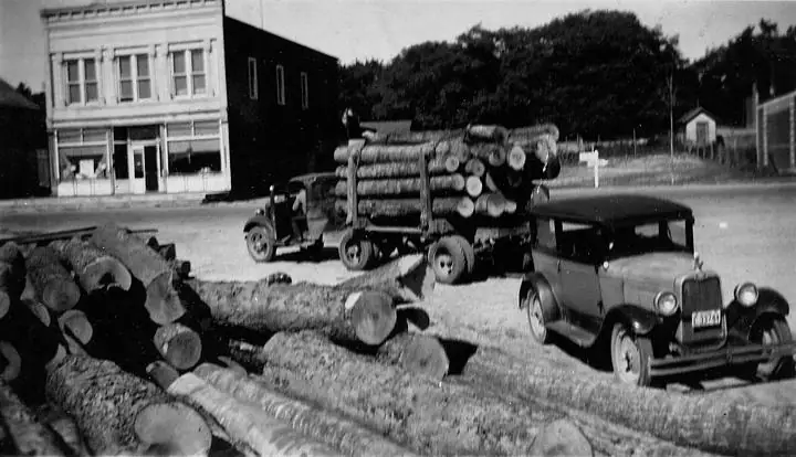A loaded logging truck, parked automobile, and piles of cut logs beside a storefront.