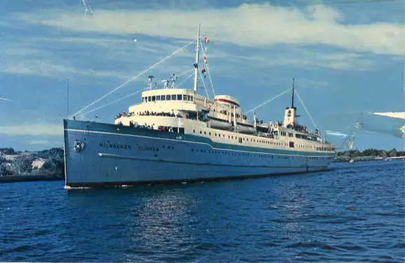 A large white-and-blue passenger ferry travels past a wooded shoreline under a partly cloudy sky.
