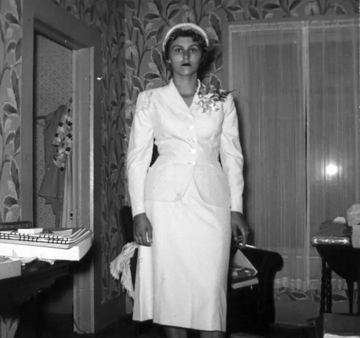 Black-and-white portrait of a woman in a white suit and hat standing indoors beside a desk and typewriter.