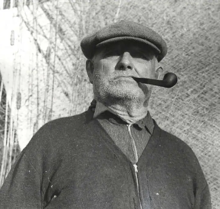 Black-and-white portrait of a bearded man in a flat cap smoking a pipe.