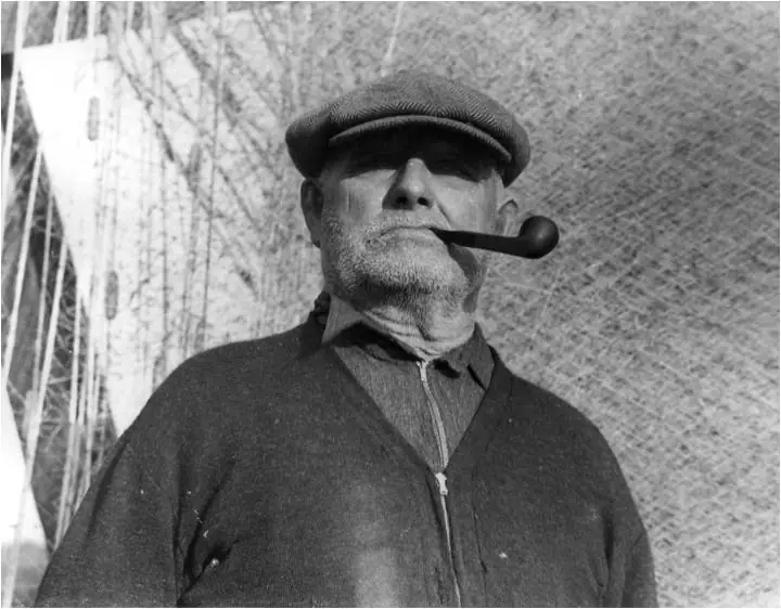 Black-and-white portrait of a bearded man wearing a flat cap and holding a curved pipe.