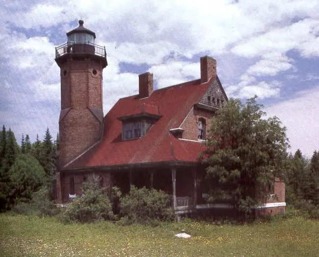 Brick lighthouse and attached red-roofed dwelling beside trees and a grassy foreground.