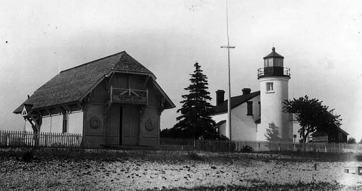 Black-and-white photograph of a lighthouse, a fenced wooden outbuilding, and a rocky shoreline.