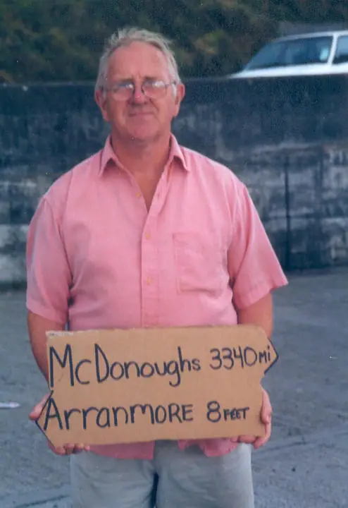 A man in a pink shirt holds a handwritten cardboard sign with distances to McDonoughs and Arranmore.