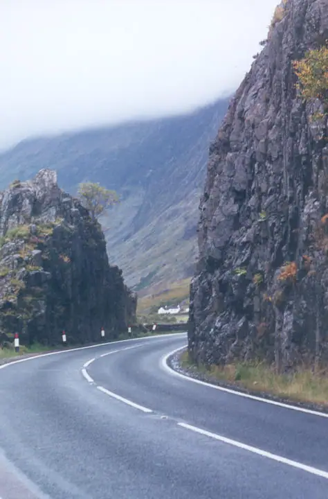 A wet paved road curves between steep rocky cliffs beneath mist-covered mountain slopes.