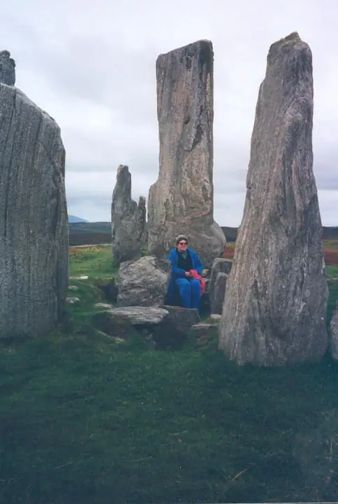 A seated woman in a blue coat among tall weathered standing stones beneath a cloudy sky.