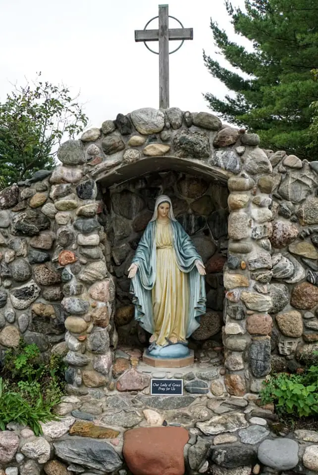 A statue of the Virgin Mary stands inside a stone grotto beneath a cross.