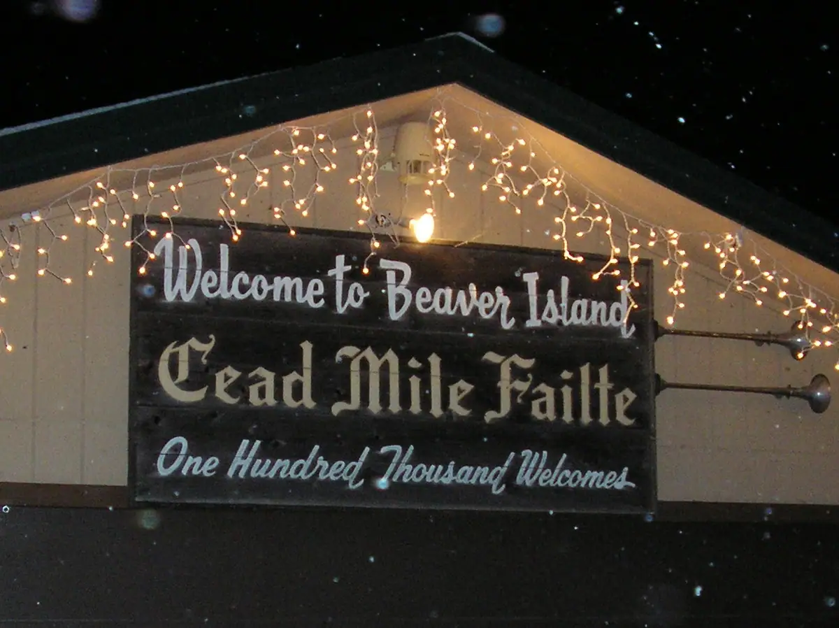 An illuminated wooden welcome sign hangs beneath strings of lights at night.