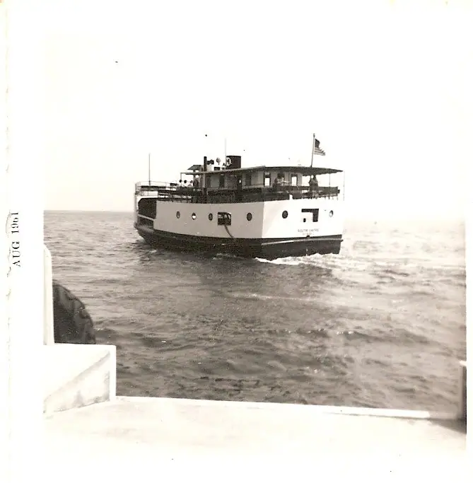 Black-and-white photograph of a ferry passing a white boat railing, with people on deck and a flag flying.