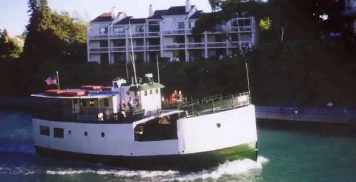 White passenger ferry passing a wooded waterfront with a large balcony-fronted building.