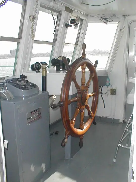 A polished wooden ship's wheel and navigation controls fill the interior of a vessel's pilothouse.