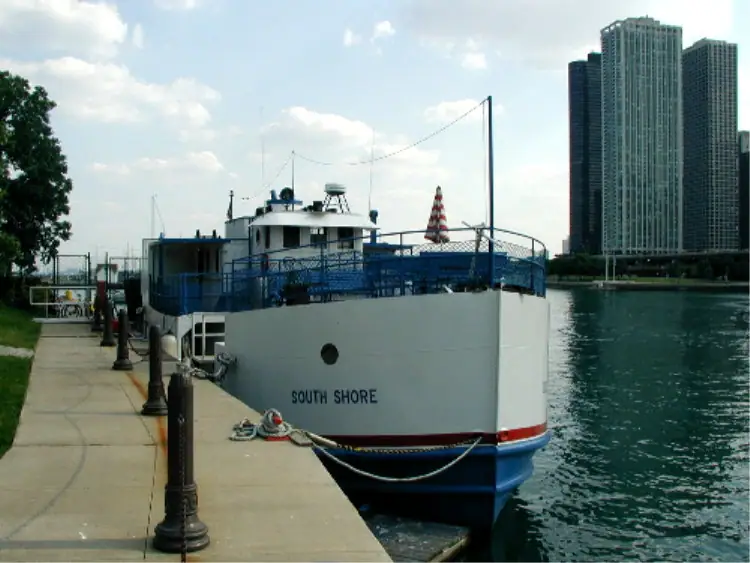 White passenger ferry moored beside a waterfront walkway and high-rise buildings.