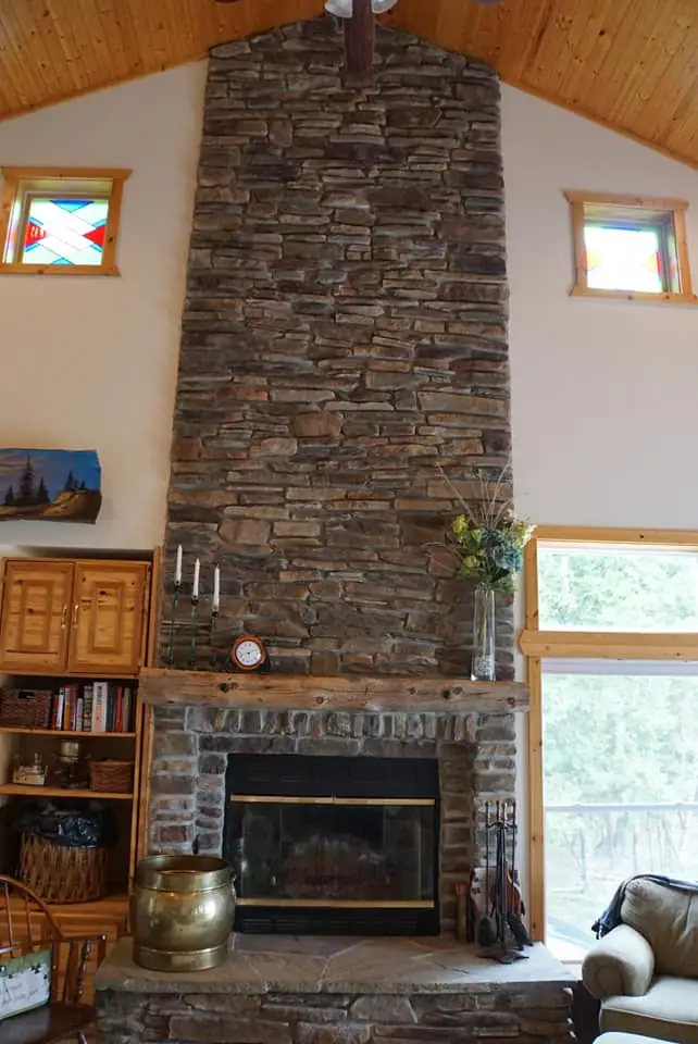 Interior view of a stone fireplace beneath a wood-paneled ceiling, with stained-glass windows and furnishings.