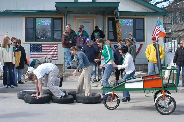 People take part in a tire obstacle course in front of a restaurant and pub, beside a small wheeled cart.